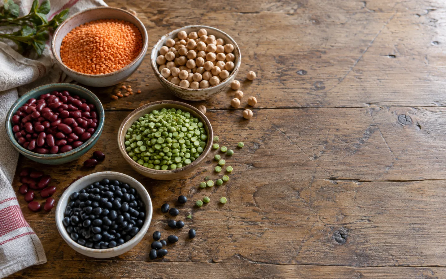 Small bowls of dried red lentils, chickpeas, kidney beans, green split peas and black beans on a wooden table in soft daylight